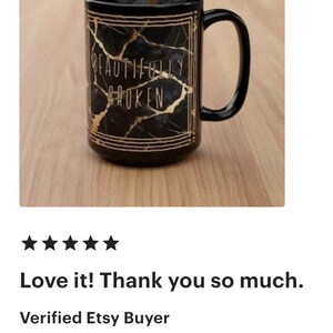 May include: Black ceramic mug with a gold marble design and the words "BEAUTIFULLY BROKEN". The mug has a black handle and is on a wooden surface. A dark liquid is being poured into the mug.
