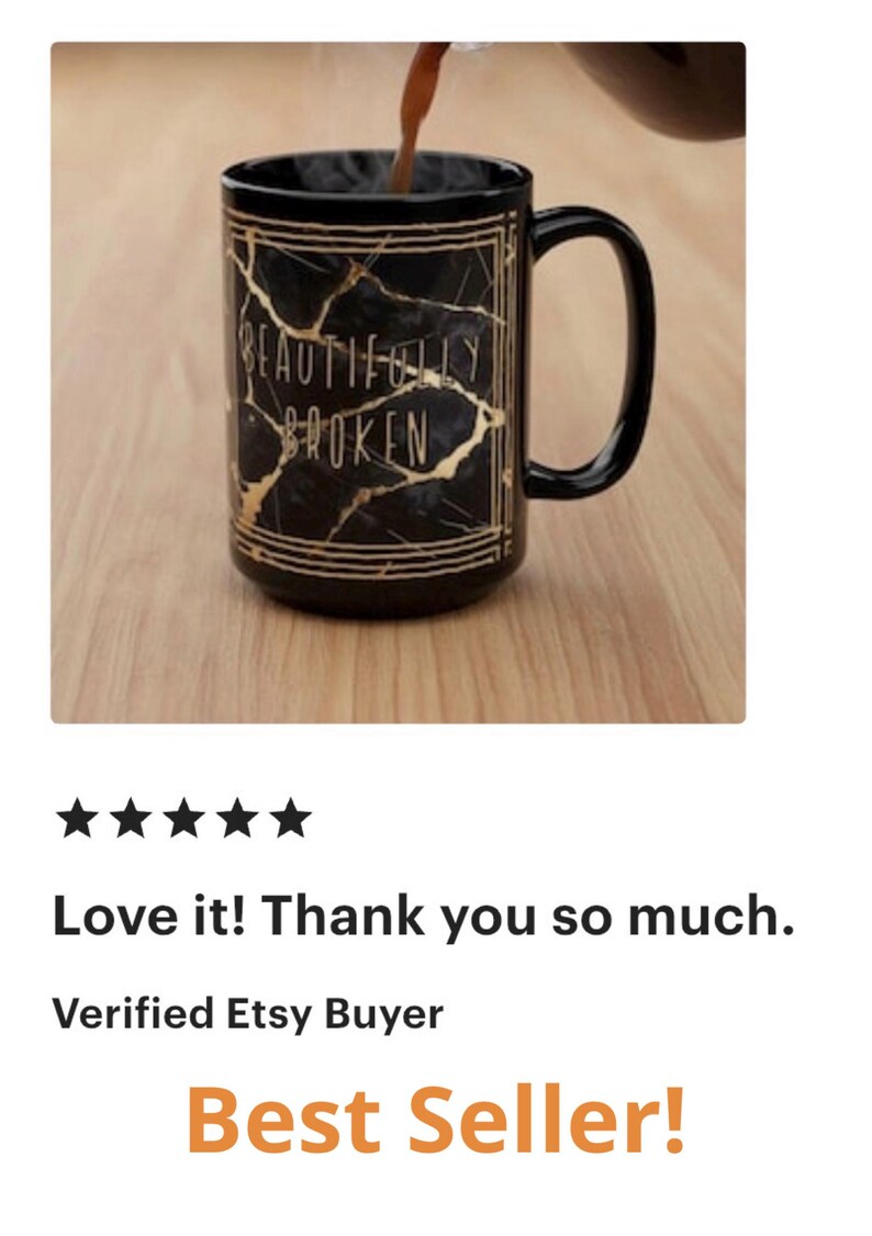 May include: Black ceramic mug with a gold marble design and the words "BEAUTIFULLY BROKEN". The mug has a black handle and is on a wooden surface. A dark liquid is being poured into the mug.