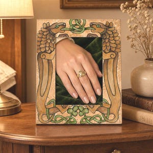 May include: A decorative picture frame with an ornate design featuring two dragon-like creatures and Celtic knotwork in green and brown. A hand wearing a gold ring is visible inside the frame.