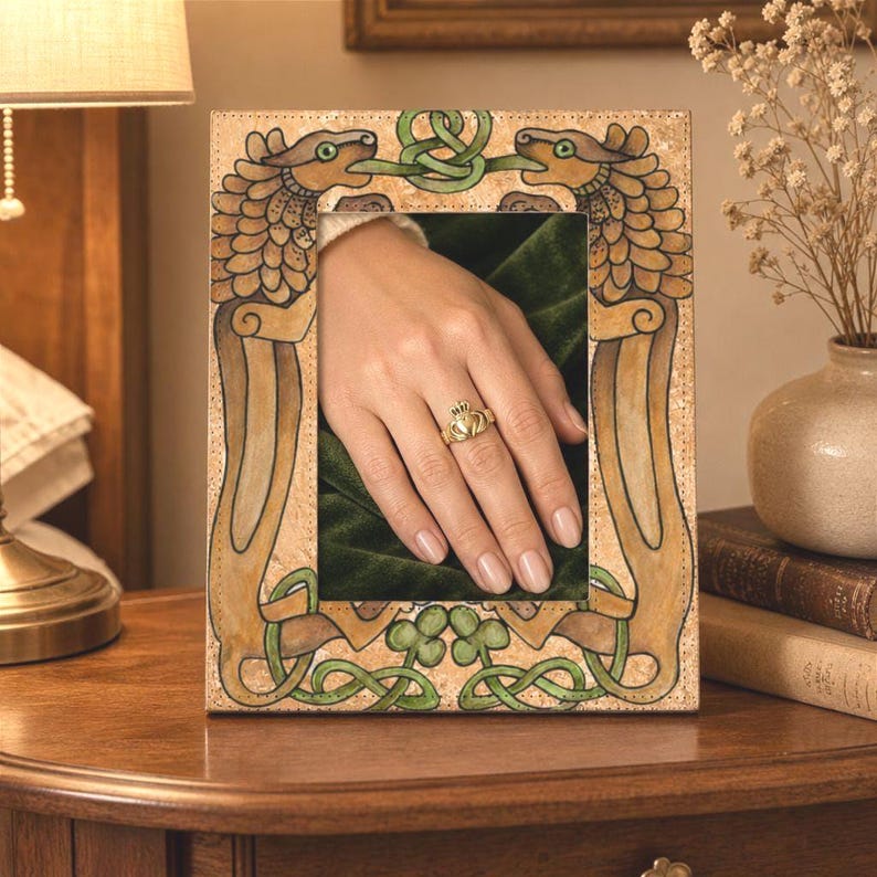 May include: A decorative picture frame with an ornate design featuring two dragon-like creatures and Celtic knotwork in green and brown. A hand wearing a gold ring is visible inside the frame.