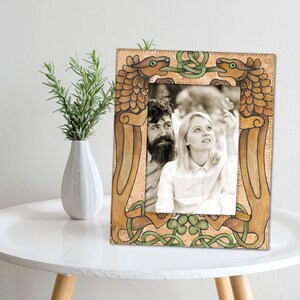 May include: A rectangular photo frame with a black and white photo of a couple. The frame is tan with green and brown Celtic knot designs and dragon-like figures. A white vase with green sprigs sits to the left on a white table.