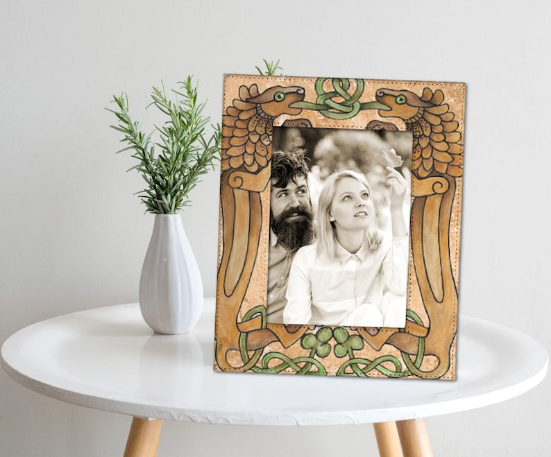 May include: A rectangular photo frame with a black and white photo of a couple. The frame is tan with green and brown Celtic knot designs and dragon-like figures. A white vase with green sprigs sits to the left on a white table.