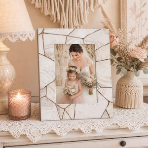 May include: A decorative photo frame with a mosaic-like design in shades of white and beige. The frame holds a photograph of a bride and a young girl, both holding bouquets. The frame is displayed on a white surface with a lace trim.