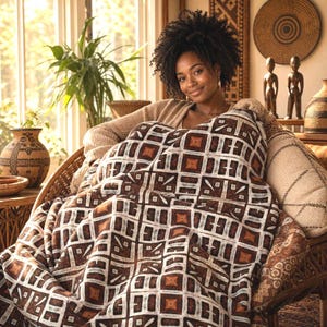 May include: A rectangular textile throw blanket featuring a repeating geometric pattern in shades of dark brown, tan, and white. The fabric displays a grid-like design with square and triangular motifs, providing a textured appearance across the surface.