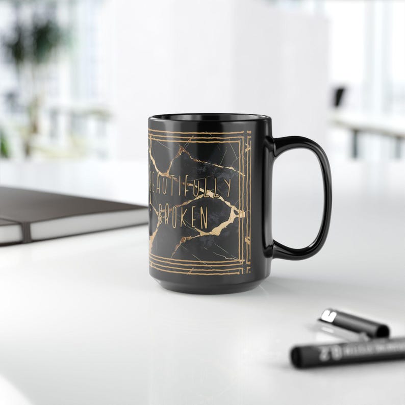 May include: A black ceramic mug with a gold and black marble design. The mug has the words "BEAUTIFULLY BROKEN" in gold lettering. The mug is on a white surface with a pen and a notebook in the background.