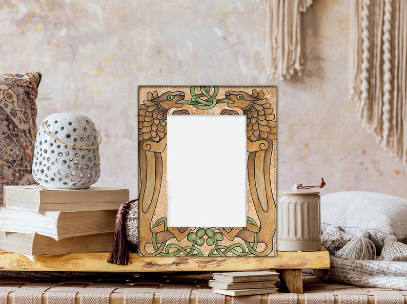 May include: A decorative picture frame with a rectangular opening. The frame features a detailed design of two stylized dragons facing each other, with green and brown accents. The frame is set on a wooden surface, with books and a decorative lantern nearby.