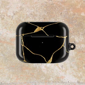 May include: Black protective case for wireless earbuds, featuring a gold vein design. The case has a glossy finish and a small loop for attaching to a keychain or lanyard. The overall design is sleek and modern.