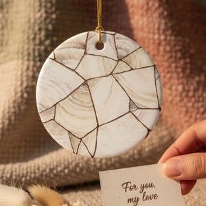 May include: A round, white ceramic ornament featuring a brown, cracked, mosaic-like pattern across its surface. The ornament hangs from a thin gold string. A small, rectangular off-white card with the text "For you, my love" is held nearby.