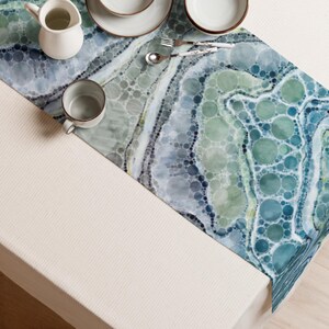 May include: A table runner featuring a pattern of overlapping circles in shades of blue, teal, and green. The runner is set with a white ceramic pitcher, a white ceramic mug, two white plates, a silver fork, and a silver spoon on a white surface.