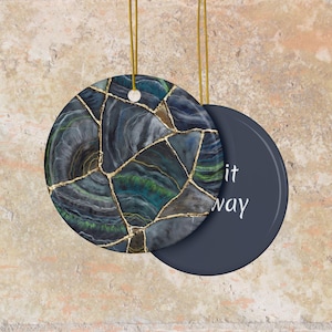 May include: Round ceramic ornament with a dark blue, green, and gold abstract design. The ornament has a gold string for hanging. The back of the ornament is dark blue with the words "it way" in white.