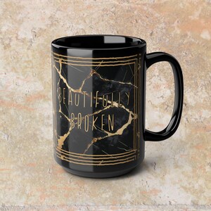 May include: Black ceramic mug with a gold geometric design and the words "BEAUTIFULLY BROKEN" in gold lettering. The mug has a black handle and a black interior. The mug is a standard size.