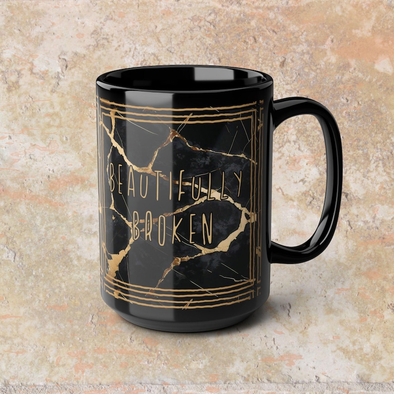 May include: Black ceramic mug with a gold geometric design and the words "BEAUTIFULLY BROKEN" in gold lettering. The mug has a black handle and a black interior. The mug is a standard size.
