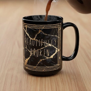 May include: A black ceramic mug with a gold cracked marble design and the words "BEAUTIFULLY BROKEN" in gold lettering. The mug has a curved handle and is being filled with a dark liquid, possibly coffee or tea.