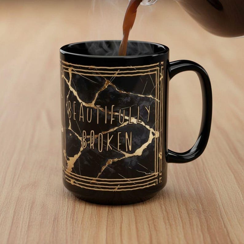 May include: A black ceramic mug with a gold cracked marble design and the words "BEAUTIFULLY BROKEN" in gold lettering. The mug has a curved handle and is being filled with a dark liquid, possibly coffee or tea.