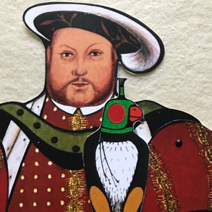 May include: A paper cutout of a man wearing a red and gold patterned robe with a white and black bird on his arm. The man is wearing a black hat with a white brim. The bird has a red and green head.