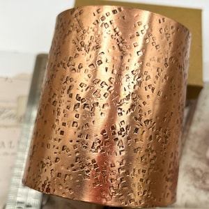 May include: A copper cuff bracelet with a hammered, textured surface. The surface is covered in a repeating pattern of small squares.