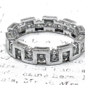 May include: A silver-colored ring with a geometric design. The ring features a series of square-shaped settings, each holding a small, clear stone. The band is marked with "925". The ring is set against a patterned background.