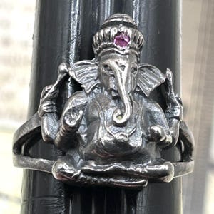 May include: A silver-toned ring featuring a detailed depiction of the Hindu deity Ganesha. The ring showcases Ganesha in a seated pose, with a prominent elephant head and multiple arms. A small, round, pink gemstone is set in the crown.