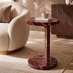 May include: A side table crafted from deep red marble with prominent white veining. The table features a circular top supported by a single vertical column that connects to a circular base, creating a distinct C-shaped profile for the furniture piece.
