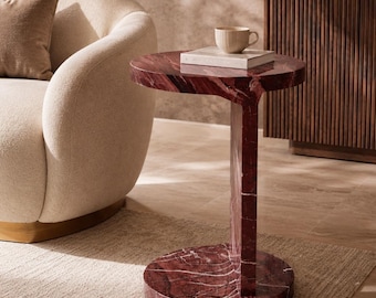 May include: A side table crafted from deep red marble with prominent white veining. The table features a circular top supported by a single vertical column that connects to a circular base, creating a distinct C-shaped profile for the furniture piece.