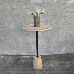May include: A small, round side table with a light-colored stone top and a black metal stem. The base is a light-colored, cone-shaped stone. A small, green vase with white flowers sits on the table. The table is approximately 24 inches tall.