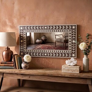 Puede incluir: Un espejo rectangular con un borde decorativo con un patrón geométrico en marrón y blanco. El espejo está montado en una pared marrón rojizo sobre una mesa de madera. Una lámpara de cobre, libros y jarrones con flores están sobre la mesa.