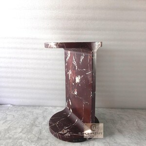 May include: A dark red marble side table featuring a circular base and a flat, overhanging top surface. The stone displays prominent white veining throughout its polished structure. A small square logo with the text CRAFTIFY DECOR is visible.