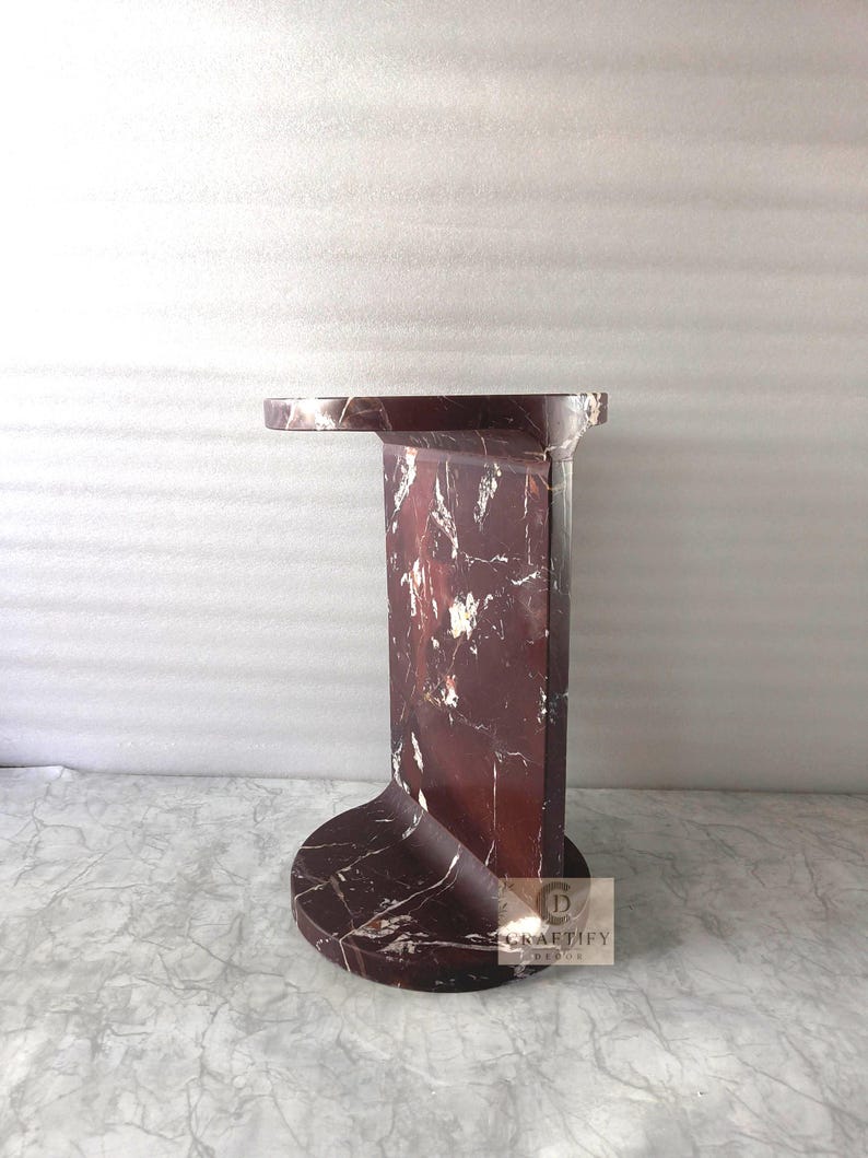 May include: A dark red marble side table featuring a circular base and a flat, overhanging top surface. The stone displays prominent white veining throughout its polished structure. A small square logo with the text CRAFTIFY DECOR is visible.