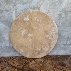 Op de afbeelding: Een ronde, beige stenen serveerplank met een textuur. De plank heeft een natuurlijk, verweerd uiterlijk met lichtere vlekken. De achtergrond is een grijze, gemarmerde muur.