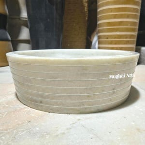 May include: A cream-coloured, round marble bowl with horizontal etched lines. The bowl has a wide, shallow design and the text "Mughal Arts" is visible on the side. The bowl is likely for decorative or functional use.