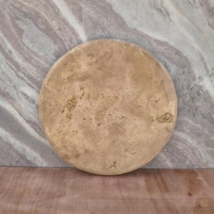 Könnte beinhalten: Ein kreisförmiges, flaches Steintablett mit einer porösen, strukturierten Oberfläche in Beige-, Hellbraun- und Sandtönen. Das Objekt weist natürliche Farbvariationen und kleine, unregelmäßige Vertiefungen auf der gesamten Vorderseite auf.