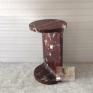 May include: A dark red marble side table with white veining, featuring a circular top and base connected by a curved vertical support. A small square label on the side displays the text CRAFTIFY DECOR with a circular logo icon above the brand name.