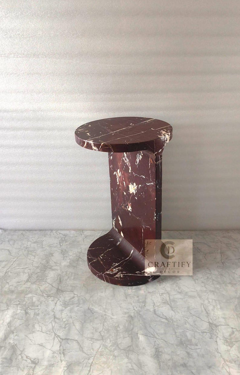 May include: A dark red marble side table with white veining, featuring a circular top and base connected by a curved vertical support. A small square label on the side displays the text CRAFTIFY DECOR with a circular logo icon above the brand name.