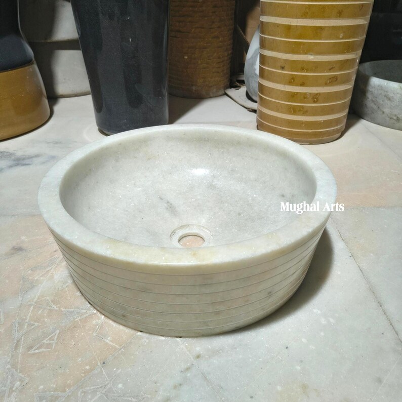 May include: A circular, off-white marble sink with a smooth interior and a textured, horizontally-lined exterior. The sink has a central drain hole and the text "Mughal Arts" is visible on the side. The sink is set on a light-coloured surface.