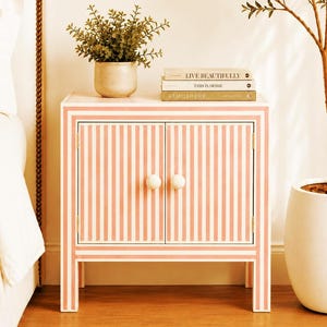 May include: A small, striped cabinet in a bedroom setting. The cabinet is painted with vertical pink and white stripes and has two doors with white, round knobs. A potted plant and stacked books sit on top.