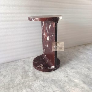May include: A dark red marble side table with white veining, featuring a circular top and base connected by a vertical central support. A square logo with the text CRAFTIFY DECOR is visible on the side of the central support structure.