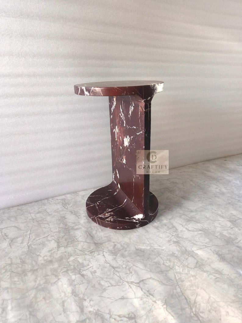 May include: A dark red marble side table with white veining, featuring a circular top and base connected by a vertical central support. A square logo with the text CRAFTIFY DECOR is visible on the side of the central support structure.