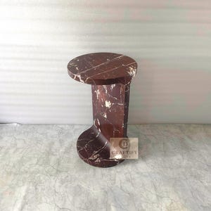 May include: A dark reddish-brown marble side table with white veining, featuring a circular top and a curved, C-shaped base. A small square logo with the text CRAFTIFY DECOR is visible on the side of the base of this stone furniture piece.