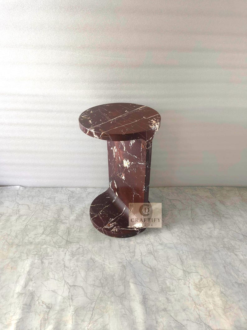 May include: A dark reddish-brown marble side table with white veining, featuring a circular top and a curved, C-shaped base. A small square logo with the text CRAFTIFY DECOR is visible on the side of the base of this stone furniture piece.