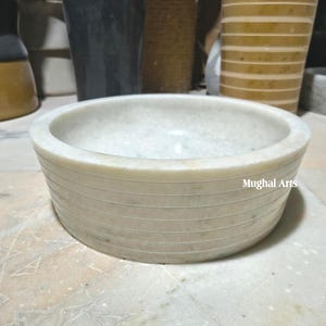 May include: A circular, off-white marble bowl with horizontal etched lines. The bowl has a smooth interior and a textured exterior. The text "Mughal Arts" is visible on the side. This decorative bowl is suitable for home decor.