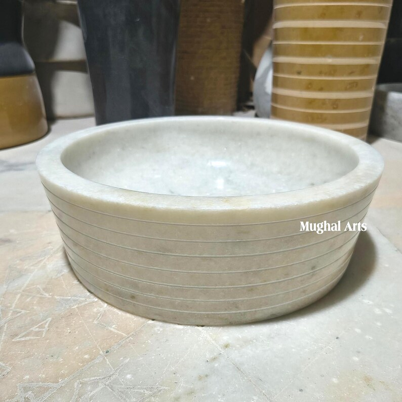 May include: A circular, off-white marble bowl with horizontal etched lines. The bowl has a smooth interior and a textured exterior. The text "Mughal Arts" is visible on the side. This decorative bowl is suitable for home decor.