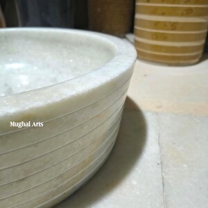 May include: A light-coloured, round marble bowl with horizontal etched lines. The bowl has a smooth, polished surface and the text "Mughal Arts" is visible on the side. The bowl is partially visible, with a blurred background.