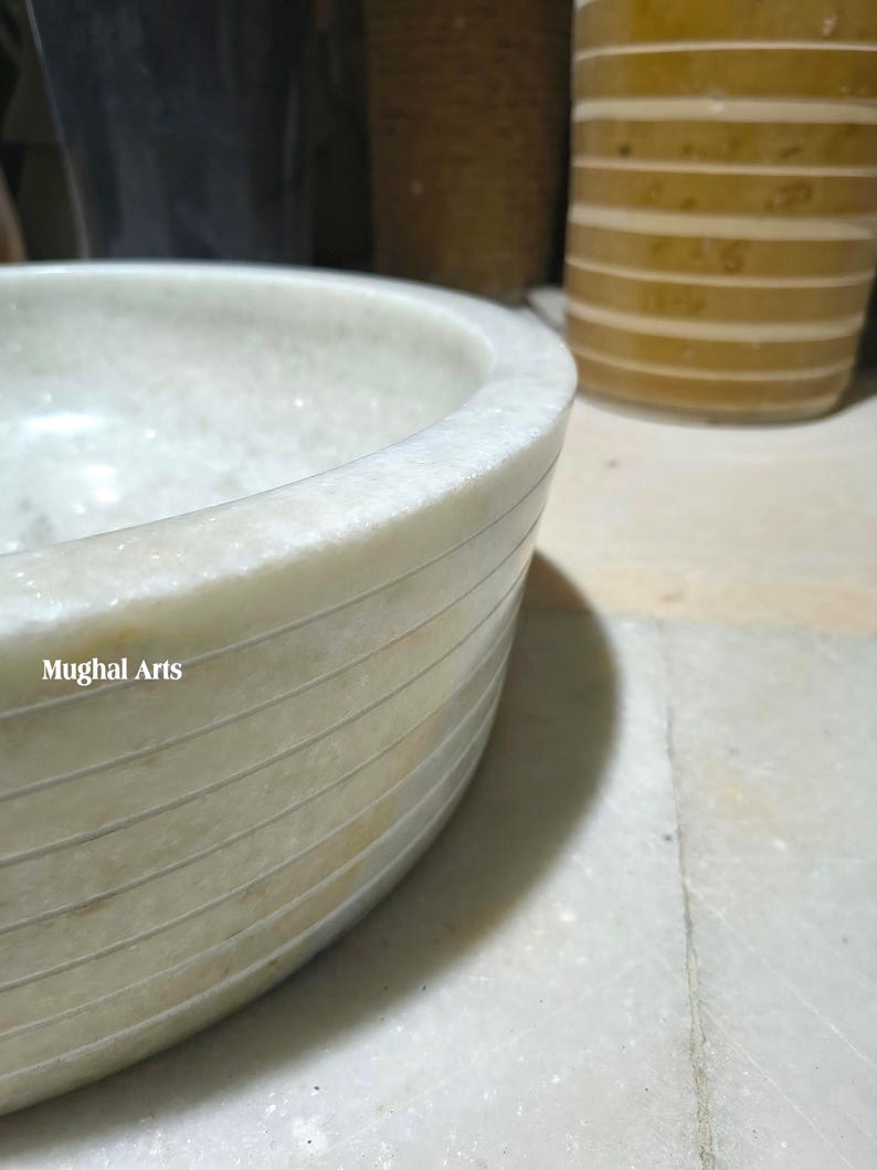 May include: A light-coloured, round marble bowl with horizontal etched lines. The bowl has a smooth, polished surface and the text "Mughal Arts" is visible on the side. The bowl is partially visible, with a blurred background.