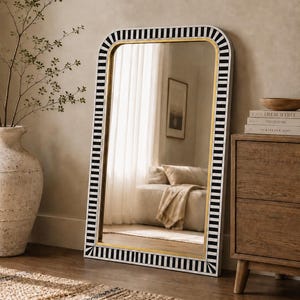 May include: A rectangular floor mirror with a black and white striped border and a gold inner frame. The mirror is leaning against a wall, reflecting a room with a couch and window. A wooden dresser and a vase with branches are also visible.