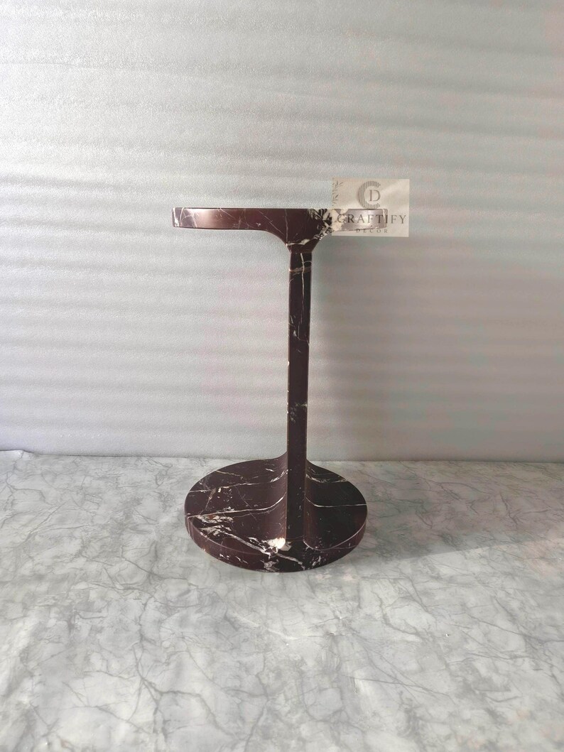 May include: A dark maroon marble side table with white veining, featuring a circular base, a vertical support column, and a round top. A small rectangular tag attached to the top edge displays the text CRAFTIFY DECOR with a circular logo.