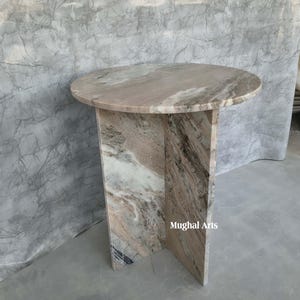 May include: A circular marble table with a light brown and grey pattern. The table has a single pedestal base. The text "Mughal Arts" is visible on the side of the base.