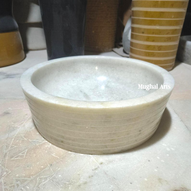 May include: A cream-coloured, round marble bowl with horizontal etched lines around the exterior. The bowl has a smooth interior and the text "Mughal Arts" is visible on the side.