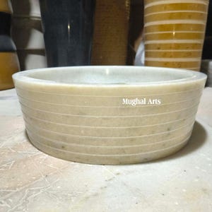 May include: A cream-coloured, round marble bowl with horizontal etched lines. The bowl has a wide, shallow design and the text "Mughal Arts" is visible on the side. The bowl is likely used for decorative or functional purposes.