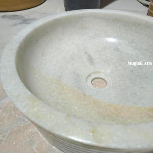 May include: A circular, off-white marble sink with a central drain hole. The sink has a smooth, polished interior and a textured exterior. The words "Mughal Arts" are visible on the right side of the sink.