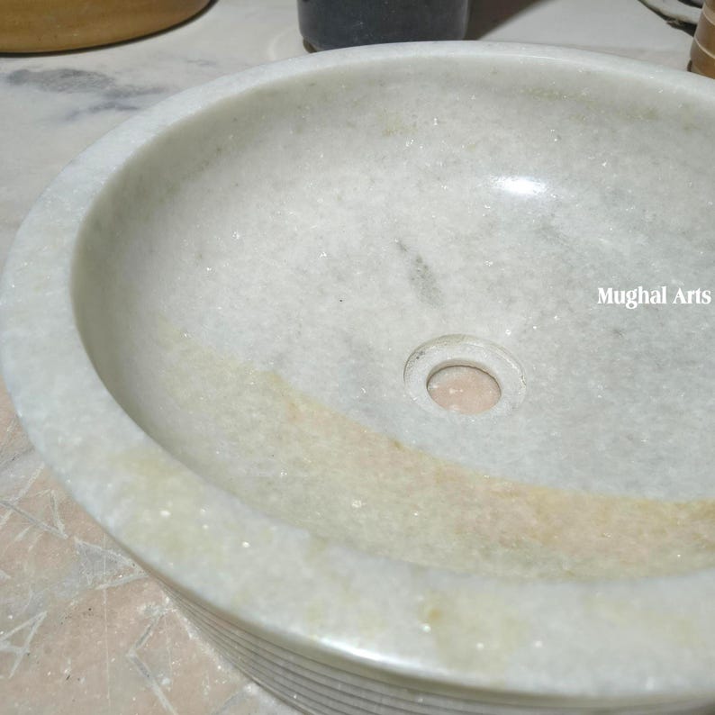 May include: A circular, off-white marble sink with a central drain hole. The sink has a smooth, polished interior and a textured exterior. The words "Mughal Arts" are visible on the right side of the sink.